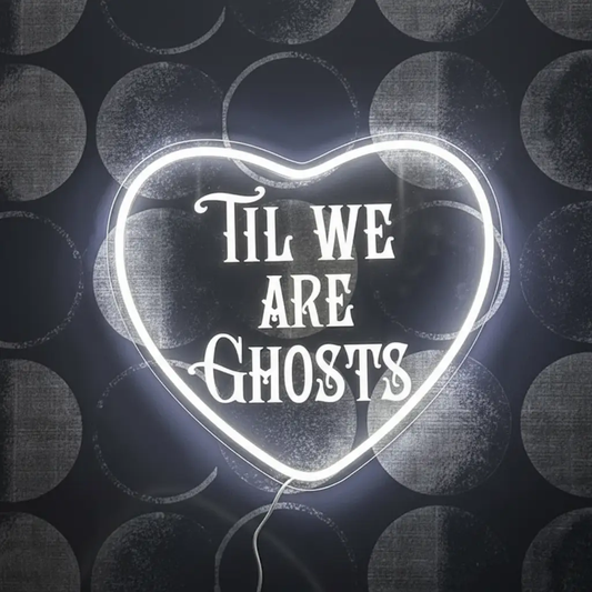 Gothic wedding neon sign reading Til We Are Ghosts in white inside a heart outline on a dark grey dotted wall