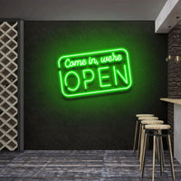Come In We're Open neon sign glowing green above bar stools
